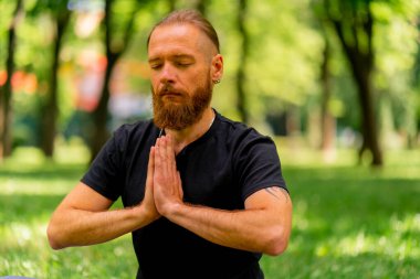 Yakışıklı sakallı adam parkta meditasyon yapıyor. Lotus pozisyonunda oturuyor. Yoga yapıyor.