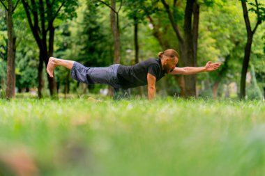 Yakışıklı sakallı adam parkta meditasyon yapıyor yoga yapıyor esneme hareketleri yapıyor ve farklı kas grupları çalıştırıyor.