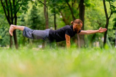 Yakışıklı sakallı adam parkta meditasyon yapıyor yoga yapıyor esneme hareketleri yapıyor ve farklı kas grupları çalıştırıyor.