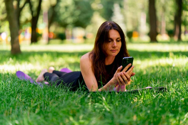 Eğitimden önce ya da sonra parkta dinlenen aktif, güzel bir Yogi kadını spor minderinde yatarken akıllı telefon görüşmesiyle konuşuyor.