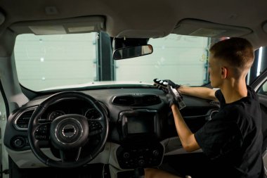 Detaylı işlemler sırasında lüks bir arabanın gösterge panelini temizlemek için sprey tabanca ve otomatik kimyasallar kullanan bir oto yıkama işçisinin yakın çekimi.