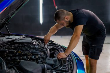 Elinde ingiliz anahtarı olan bir araba tamircisi kaputu açık, zincir renkli bir filmde araba tamir ediyor.