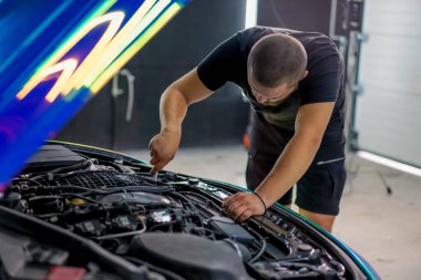 Elinde ingiliz anahtarı olan bir araba tamircisi kaputu açık, zincir renkli bir filmde araba tamir ediyor.