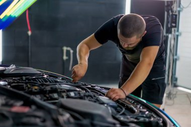 Elinde ingiliz anahtarı olan bir araba tamircisi kaputu açık, zincir renkli bir filmde araba tamir ediyor.