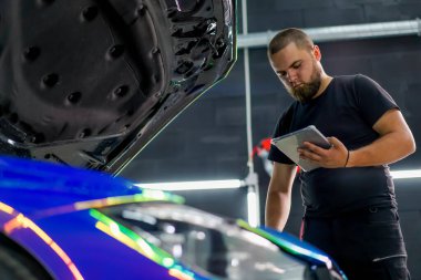 Kaputu açık pahalı bir arabanın yanında dururken araba servisi çalışanı elindeki tablete bakıyor.