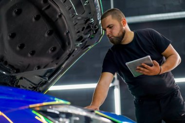 Kaputu açık pahalı bir arabanın yanında dururken araba servisi çalışanı elindeki tablete bakıyor.
