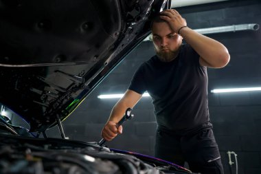 Elinde İngiliz anahtarı olan bir araba tamircisi kafasını tutuyor ve kaputu açık bir şekilde arabayı tamir etmeye çalışıyor.