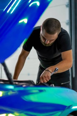 Elinde ingiliz anahtarı olan bir araba tamircisi kaputu açık, zincir renkli bir filmde araba tamir ediyor.