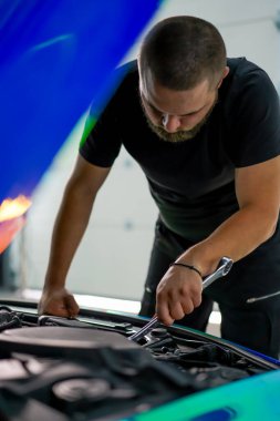 Elinde ingiliz anahtarı olan bir araba tamircisi kaputu açık, zincir renkli bir filmde araba tamir ediyor.