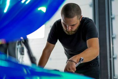 Elinde ingiliz anahtarı olan bir araba tamircisi kaputu açık, zincir renkli bir filmde araba tamir ediyor.