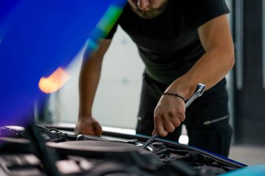 Elinde ingiliz anahtarı olan bir araba tamircisi kaputu açık, zincir renkli bir filmde araba tamir ediyor.