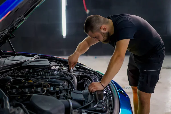 Elinde ingiliz anahtarı olan bir araba tamircisi kaputu açık, zincir renkli bir filmde araba tamir ediyor.