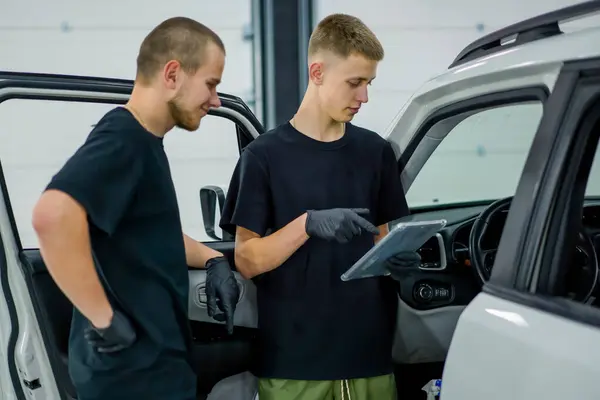 two car mechanics holding a tablet with an open green screen next to the open door of white car