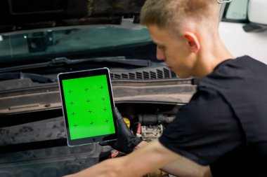 İki oto tamircisi kaputu açık beyaz SUV 'nin yanında duruyor ve yeşil ekranlı bir tablete bakın.