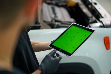 İki oto tamircisi kaputu açık beyaz SUV 'nin yanında duruyor ve yeşil ekranlı bir tablete bakın.