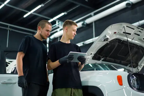 Two auto mechanics stand next to white SUV with the hood open and look at the tablet they hold in their hands while repairing the car