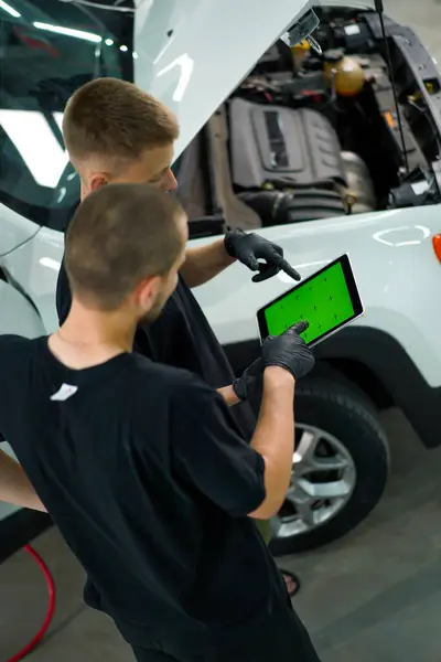Two auto mechanics stand next to white SUV with the hood open and look at a tablet with a green screen open while repairing the car
