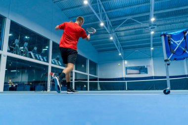 Genç adam tenis kortunda tenis antrenörlüğü yaparken raketle topa vuruyor.