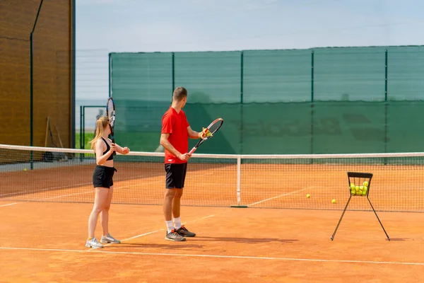 Genç bir koç, acemi bir kıza tenis oynamayı öğretir, tenis kortunda tenis topuyla raket pratiği yapar.
