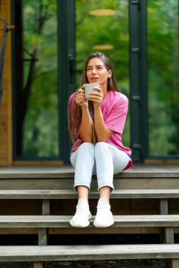 Düşünceli bir kız elinde bir fincan çayla sabah ormandaki ahşap kulübesinde oturur.