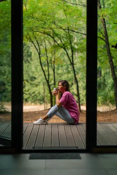 a young smiling woman sits sideways on a terrace overlooking the forest and enjoys the silence
