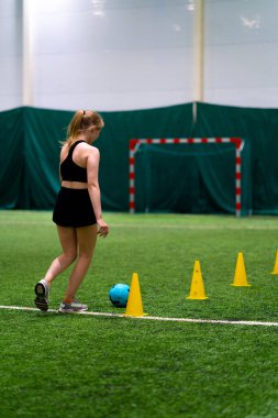 Genç kız futbol sahasında antrenman yapıyor topu cipslerin ya da konilerin arasına sürüyor ve maça hazırlanıyor.