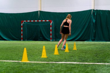 Genç kız futbol sahasında antrenman yapıyor topu cipslerin ya da konilerin arasına sürüyor ve maça hazırlanıyor.