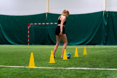 Genç kız futbol sahasında antrenman yapıyor topu cipslerin ya da konilerin arasına sürüyor ve maça hazırlanıyor.