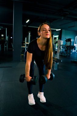 Vücut geliştirme müsabakasında ellerindeki dambıllarla egzersiz yapan konsantre fitness kadın antrenörü.