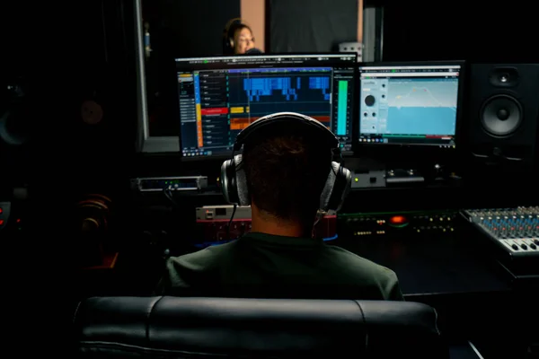a male sound engineer wearing headphones sits at the mixing desk with his back to the camera and looks at the sound tracks in the computer monitor