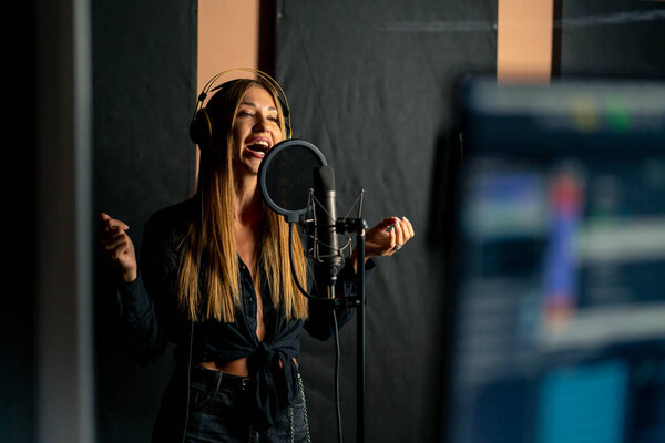 девушка певица поет в студии звукозаписи с наушниками перед микрофоном, чтобы создать новую песню