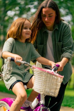 Aile portresi genç anne kızına şehir parkında bisiklet sürmeyi öğretiyor mutlu bir çocukluk ve bakım.