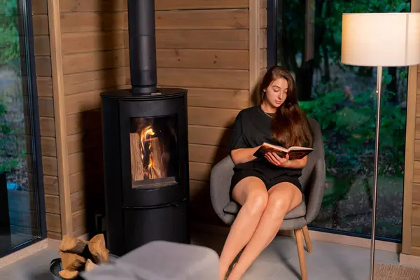 A pensive girl sits by the fireplace in the evening and carefully reads a book in a wooden house
