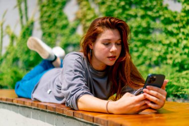 Genç bir kız elinde telefonuyla parkta bir bankta uzanıyor. Sosyal ağda bir arkadaşıyla mesajlaşıyor ya da haberleri okuyor.