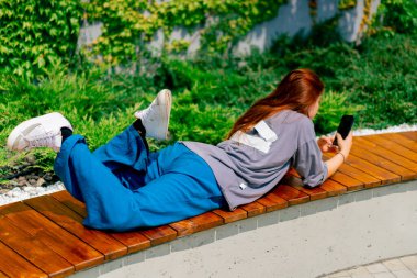 Genç bir kız elinde telefonuyla parkta bir bankta uzanıyor. Sosyal ağda bir arkadaşıyla mesajlaşıyor ya da haberleri okuyor.