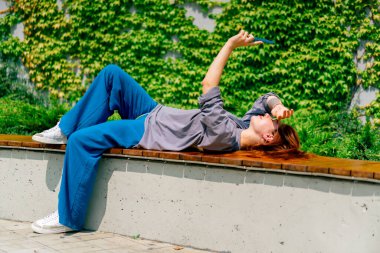 Genç bir kız elinde telefonla parkta bankta uzanmış selfie makinesinde kendi fotoğrafını çekiyor.