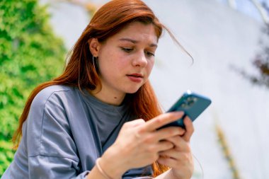 Genç bir kız parkta bir bankta elinde telefonla oturmuş sosyal ağda arkadaşıyla sohbet ediyor. Haberleri okuyor.