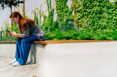 Genç bir kız parkta bir bankta elinde telefonla oturmuş sosyal ağda arkadaşıyla sohbet ediyor. Haberleri okuyor.