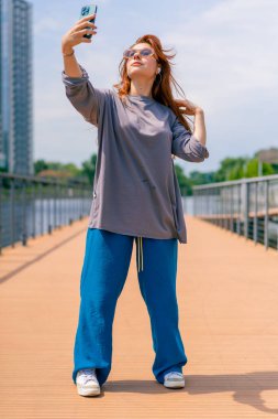 Güneş gözlüklü, elinde telefon olan genç, kızıl saçlı bir kız selfie makinesinde kendi fotoğrafını çekiyor.