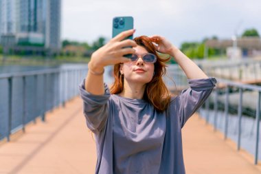 Güneş gözlüklü, elinde telefon olan genç, kızıl saçlı bir kız selfie makinesinde kendi fotoğrafını çekiyor.