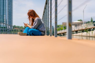 Genç kızıl saçlı bir kız bir iskelede ya da köprüde telefonla oturuyor ve sosyal ağlarda iletişim kuran bir arkadaşıyla iletişim kuruyor.