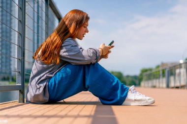 Genç kızıl saçlı bir kız bir iskelede ya da köprüde telefonla oturuyor ve sosyal ağlarda iletişim kuran bir arkadaşıyla iletişim kuruyor.