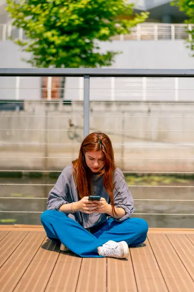 a young red-haired girl sits on a pier or bridge with phone and communicates with a friend in the messenger of communication in social networks