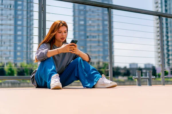 a young red-haired girl sits on a pier or bridge with phone and communicates with a friend in the messenger of communication in social networks