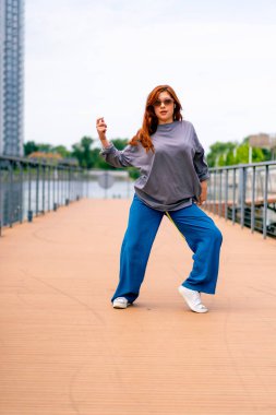 Genç, kızıl saçlı sokak hip-hop dansçısı spor kıyafetleri içinde doğaçlama danslar yapıyor. Bize briç oynuyor. Yoldan geçenlerin ilgisini çekiyor.