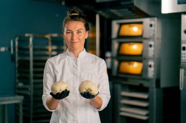 Gülümseyen bir fırıncı kadın fırının arka planında profesyonel bir mutfakta duruyor ve elinde hamur parçaları tutuyor.