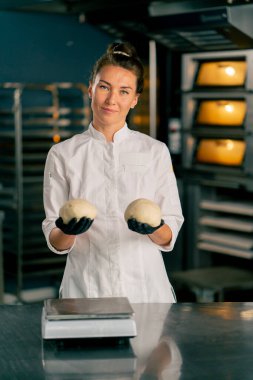 Gülümseyen bir fırıncı kadın fırının arka planında profesyonel bir mutfakta duruyor ve elinde hamur parçaları tutuyor.