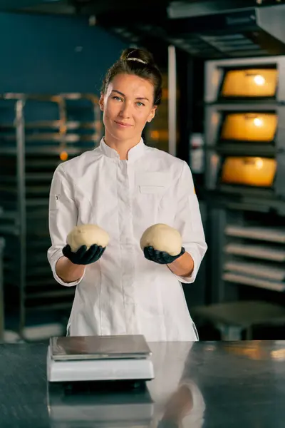 微笑的女面包师站在一个专业的厨房里 靠着烤箱 手里拿着几块面团 — 图库照片