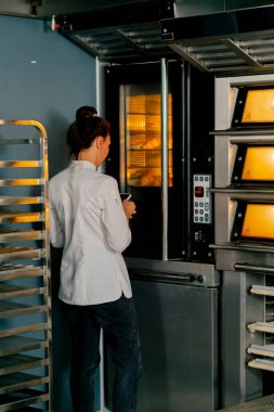 Bir fırıncı kadın fırının başında durur, fırının sıcaklığını ayarlar ve taze pişmiş ürünlerin pişirilmesini izler.
