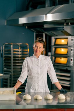 Kadın fırıncı, fırında pişirmek için hazırlanmış hamur ile profesyonel bir mutfak yüzeyinin arkasında duruyor ve kameraya bakıyor.